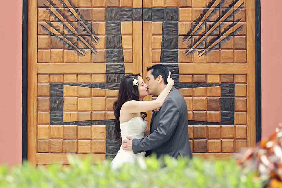Pareja-de-novios-besandose-frente-a-una-gran-puerta-de-madera-tallada-con-relieves-geometricos-fotografia-de-bodas-profesional-en-Chia
