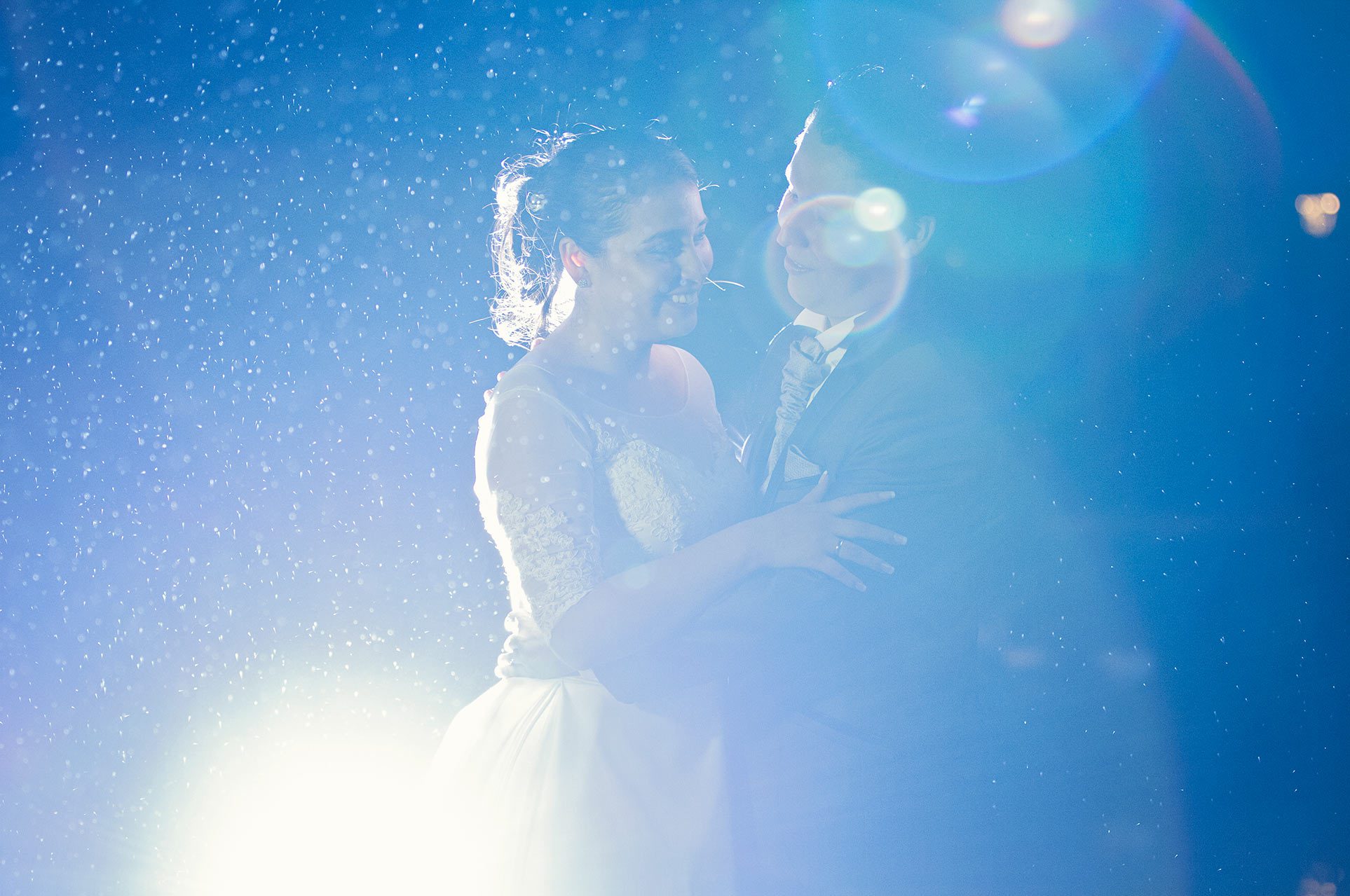 Novios abrazados en sesión de boda nocturna con luz azul, efecto bokeh y partículas brillantes, fotografía romántica y cinematográfica.