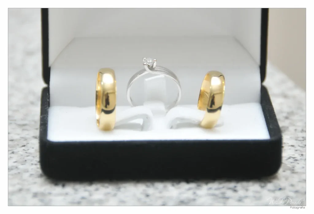 Tres anillos de boda (dos alianzas de oro amarillo y un anillo de compromiso de plata con diamante) presentados dentro de una caja de joyería negra abierta sobre una superficie de piedra.
