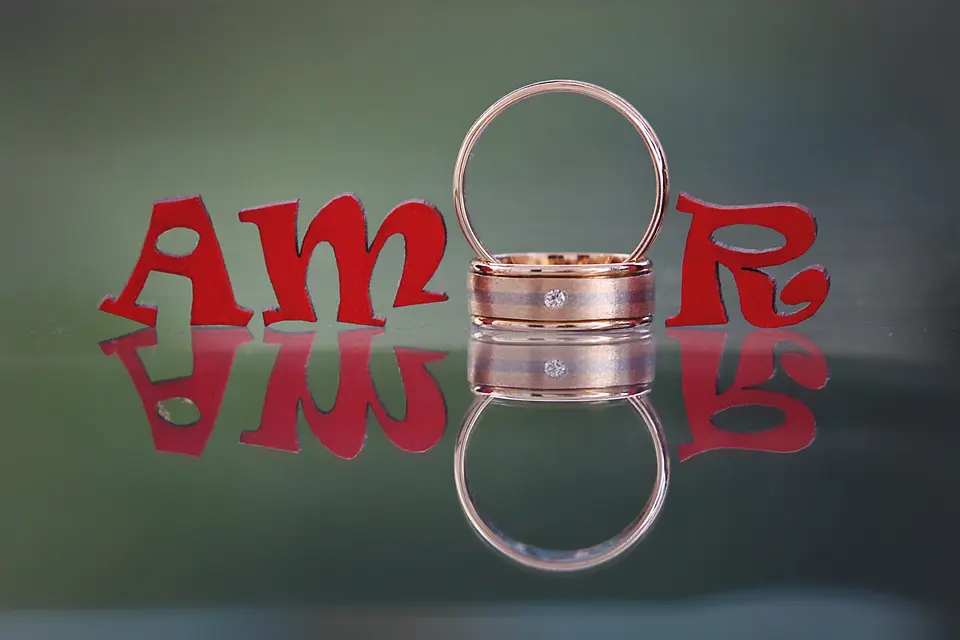 Primer plano de dos argollas de matrimonio de oro rosa colocadas entre letras decorativas rojas que forman la palabra "AMOR", con un reflejo nítido sobre una superficie de cristal. Wilder Fotografia