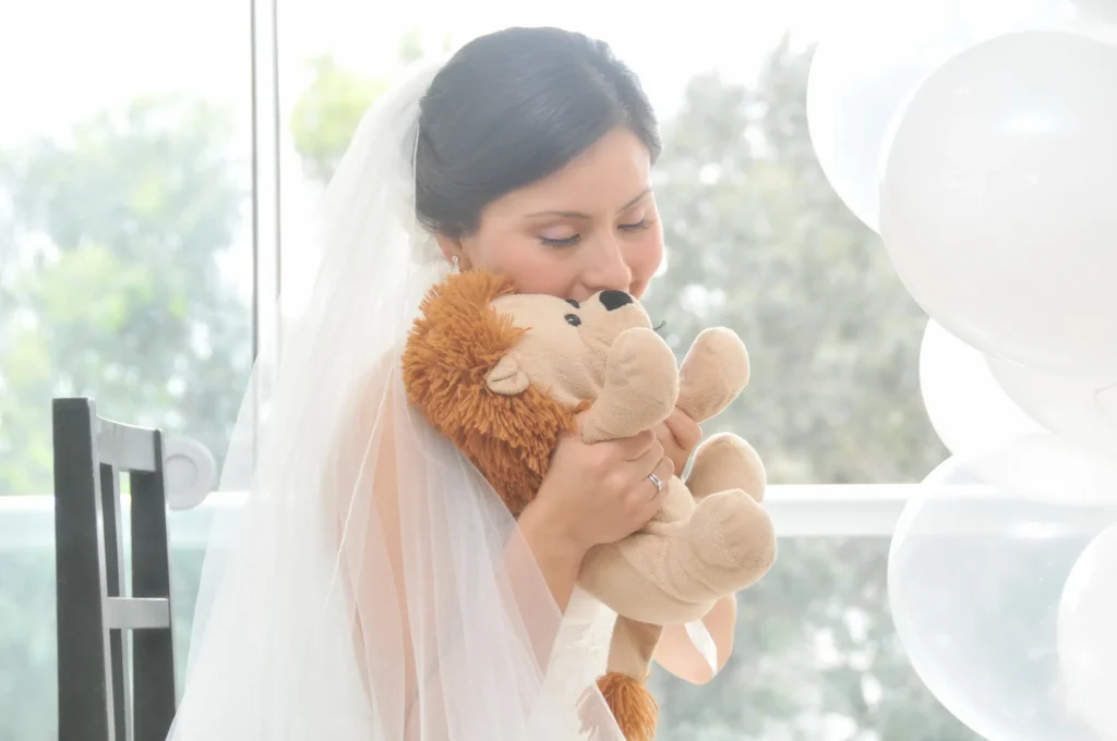 Retrato de una novia con velo cerrando los ojos y abrazando con ternura a un león de peluche, con globos blancos visibles en el fondo.