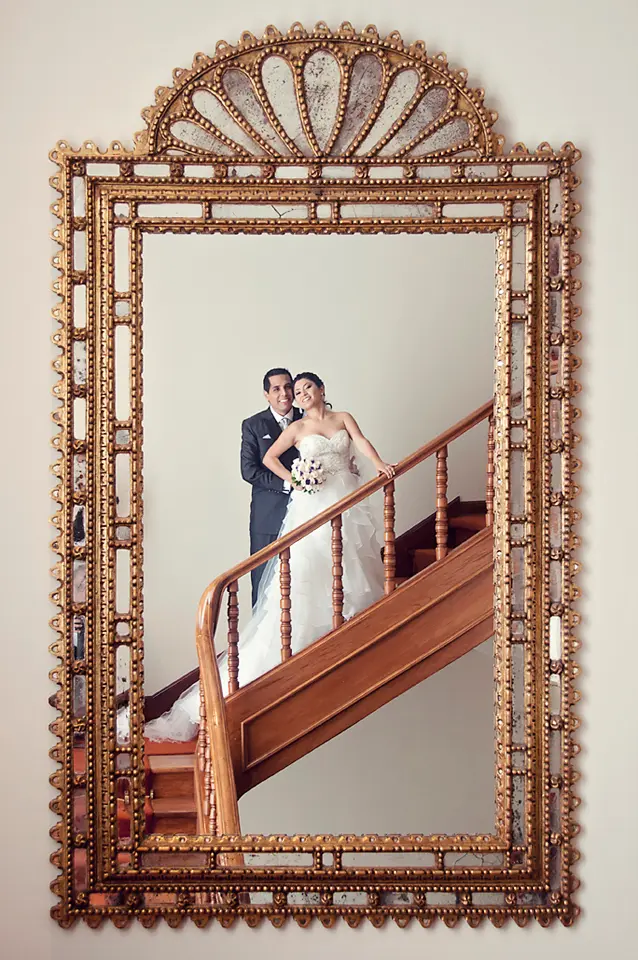 Reflejo de los novios posando en una escalera de madera visto a través de un gran espejo antiguo con marco dorado ornamentado. sesión de fotos preboda en hotel clásico.