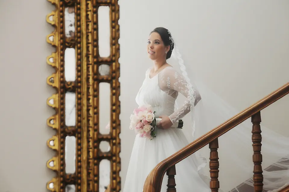 Novia sosteniendo su ramo y sonriendo en la escalera junto a un gran marco dorado antiguo, sesión fotográfica en la Sabana de Bogotá.