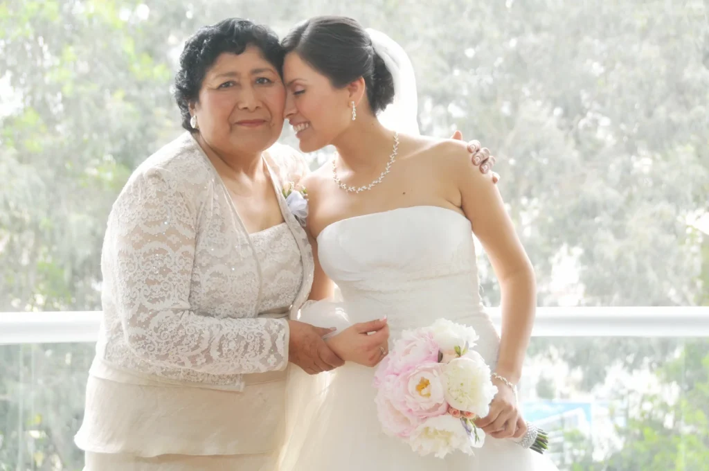 Novia con vestido de encaje blanco abrazando sonriente a su madre en un entorno luminoso.