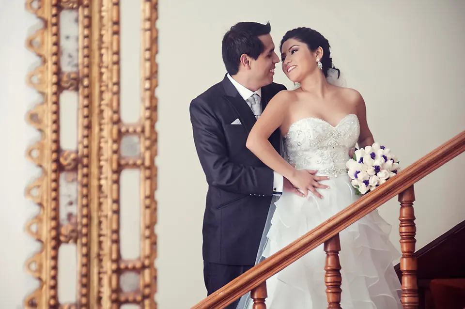 Pareja de recién casados sonriendo y posando con elegancia en una escalera de madera con pasamanos clásico, junto a un marco dorado ornamental.