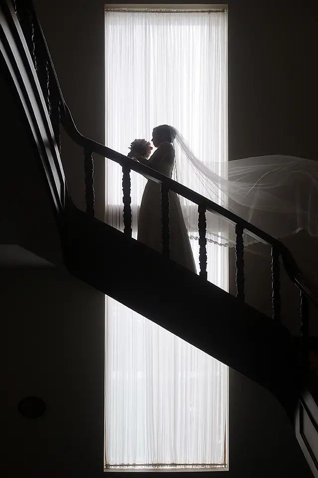 Silueta de una novia con velo largo y ramo de flores, posando en una elegante escalera de madera frente a un gran ventanal iluminado. Sesión de bodas en hacienda Cundinamarca