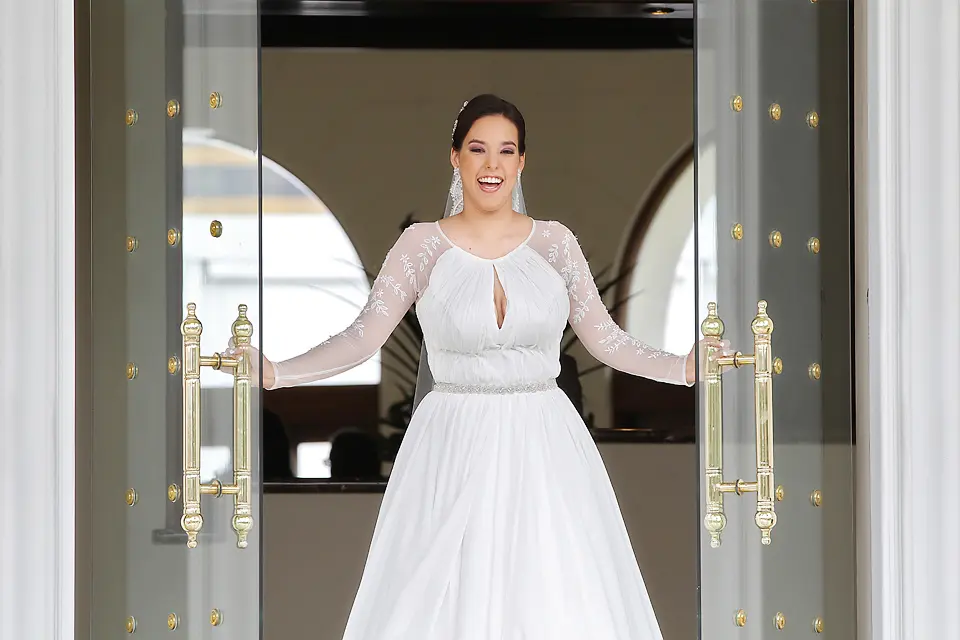 Novia feliz y sonriente abriendo unas grandes puertas de cristal con manijas doradas, mostrando su vestido de bodas con mangas de encaje.
