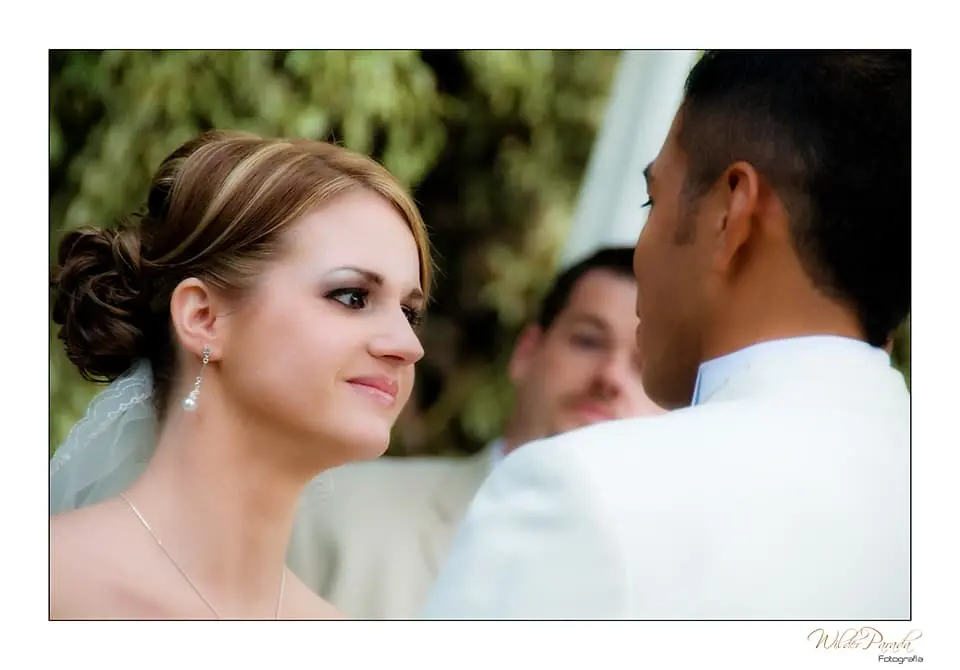 Primer plano de la novia mirando con una sonrisa sutil al novio durante la ceremonia de boda, con un invitado de fondo desenfocado.