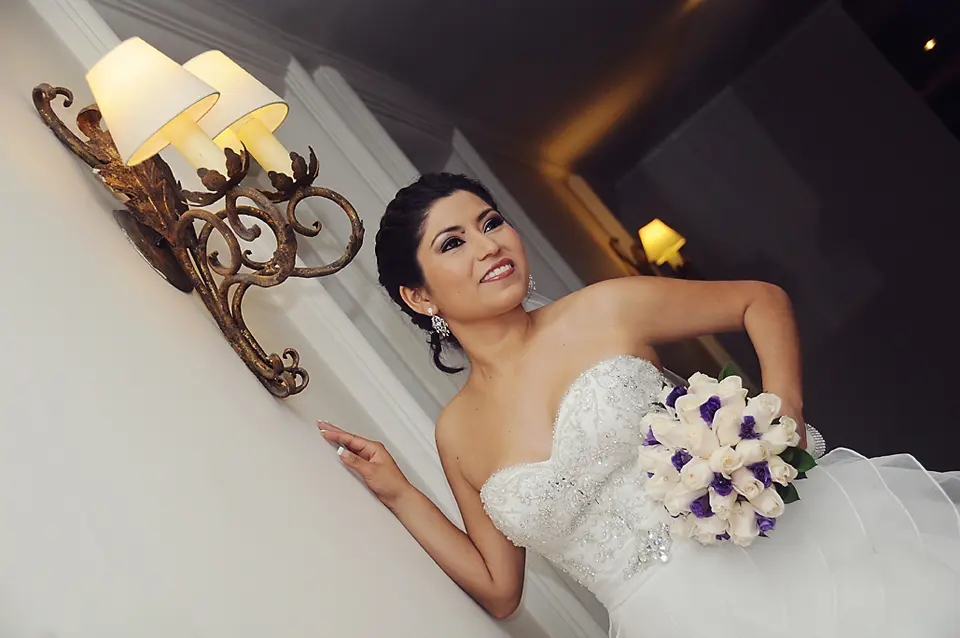 Retrato de la novia con un ramo de rosas blancas y púrpuras, posando con una mano en la pared bajo una lámpara de aplique clásica.