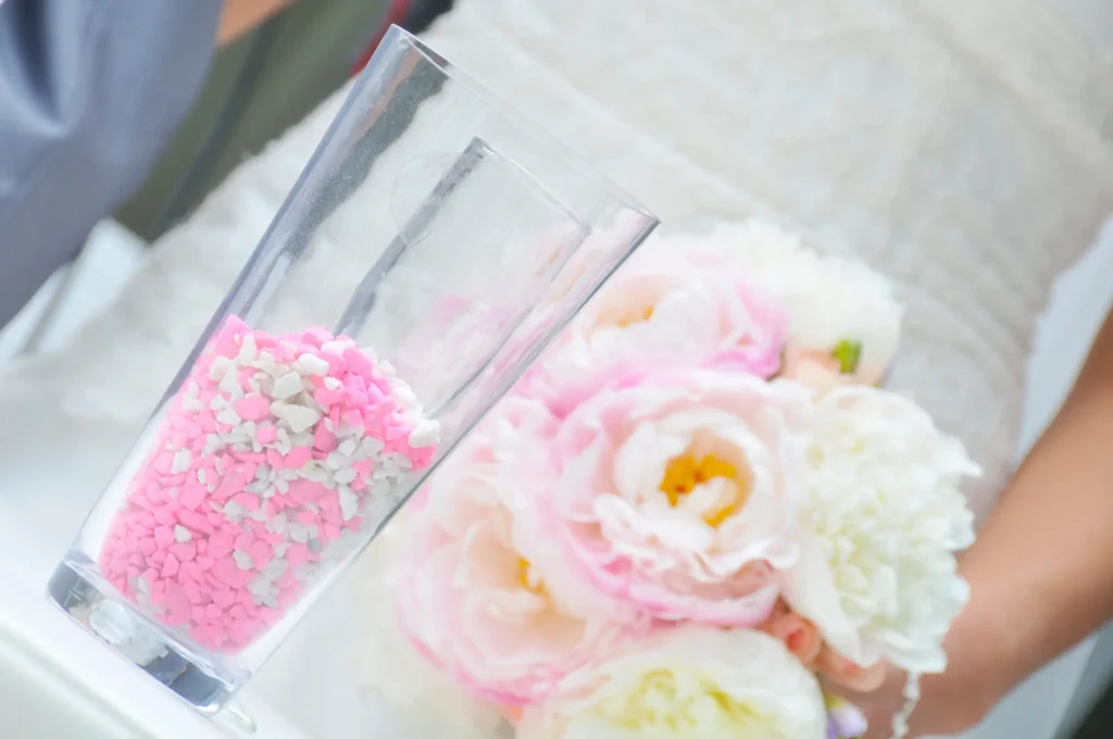 Primer plano de frasco de cristal con arena de colores y ramo de peonías blancas y rosas al fondo.