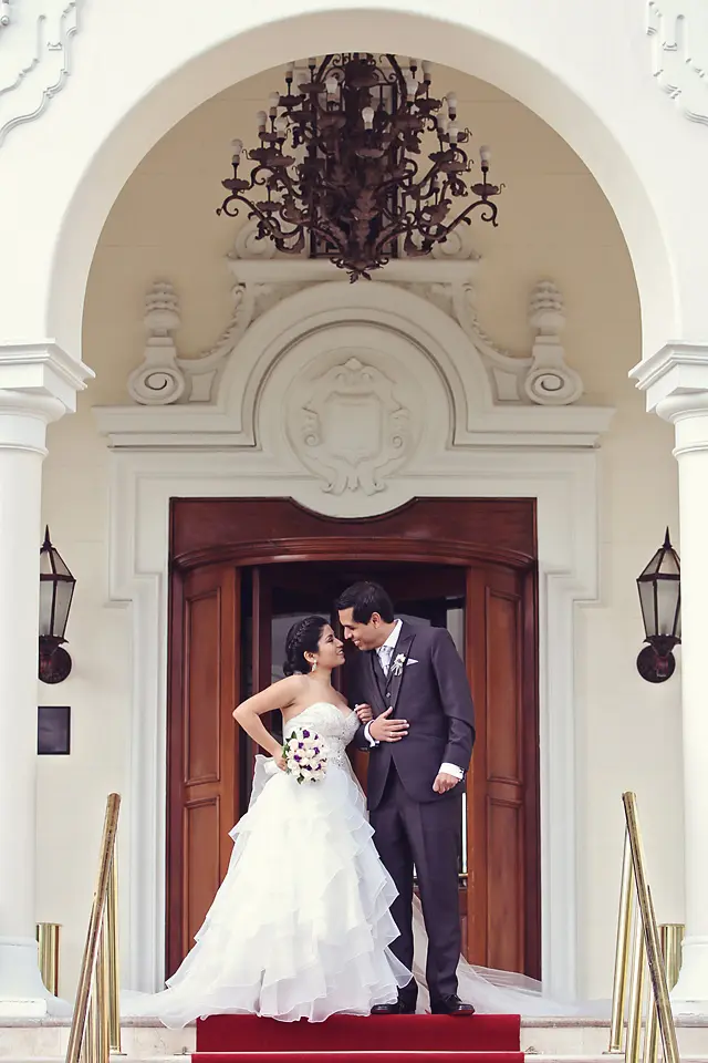 Los novios se miran con ternura en la entrada de un edificio de arquitectura clásica con grandes columnas blancas y una alfombra roja.
