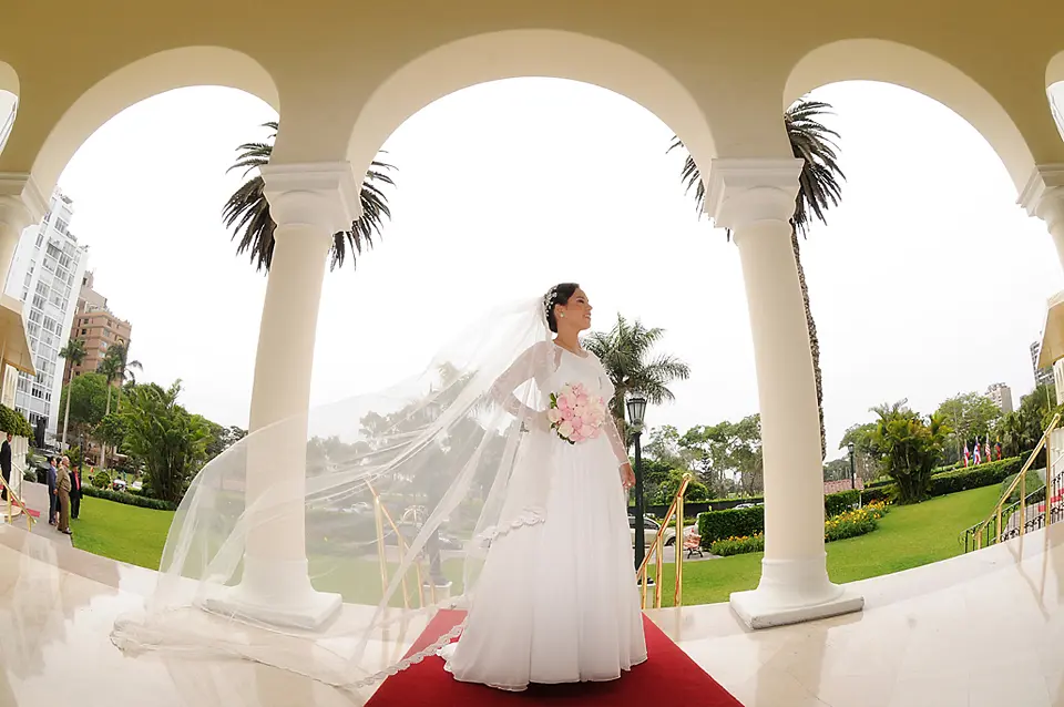 Fotografía artística de una novia con velo largo flotando al viento, posando bajo una estructura de arcos blancos y palmeras.