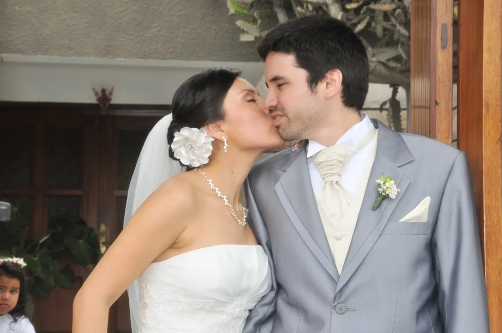 Novia besando en la mejilla al novio frente a una puerta de madera clásica.