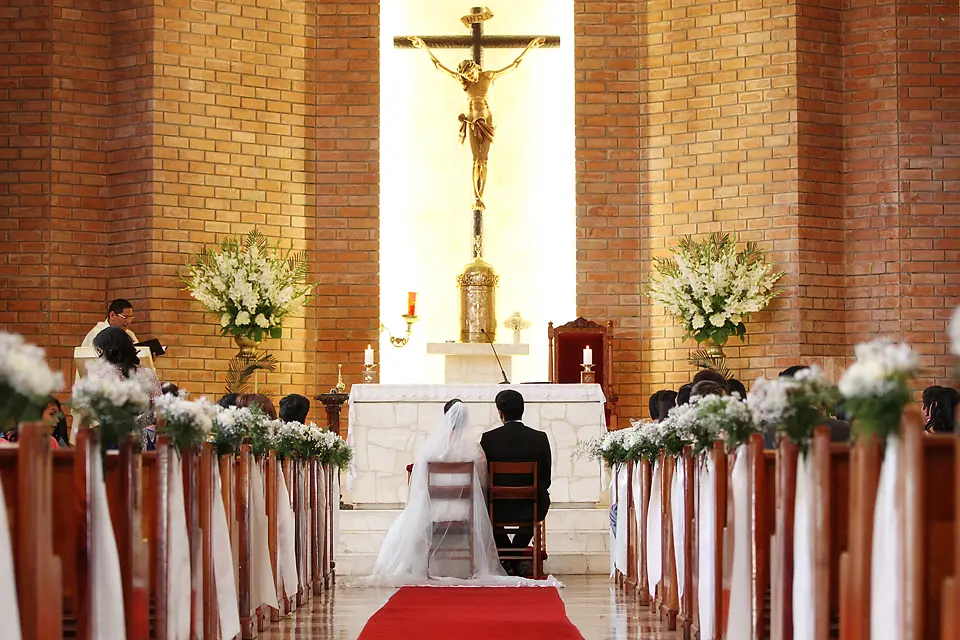 Vista desde el pasillo central de una iglesia con los novios sentados en el altar frente a un gran crucifijo dorado durante la misa de bodas