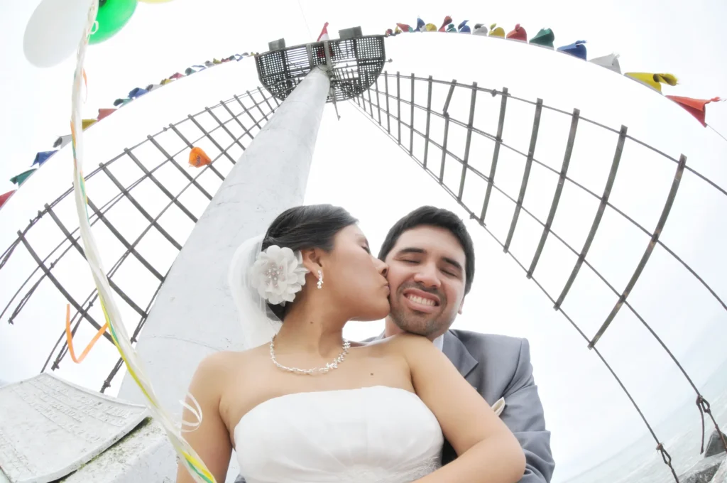 Plano contrapicado con lente ojo de pez de novios posando bajo una estructura náutica blanca.