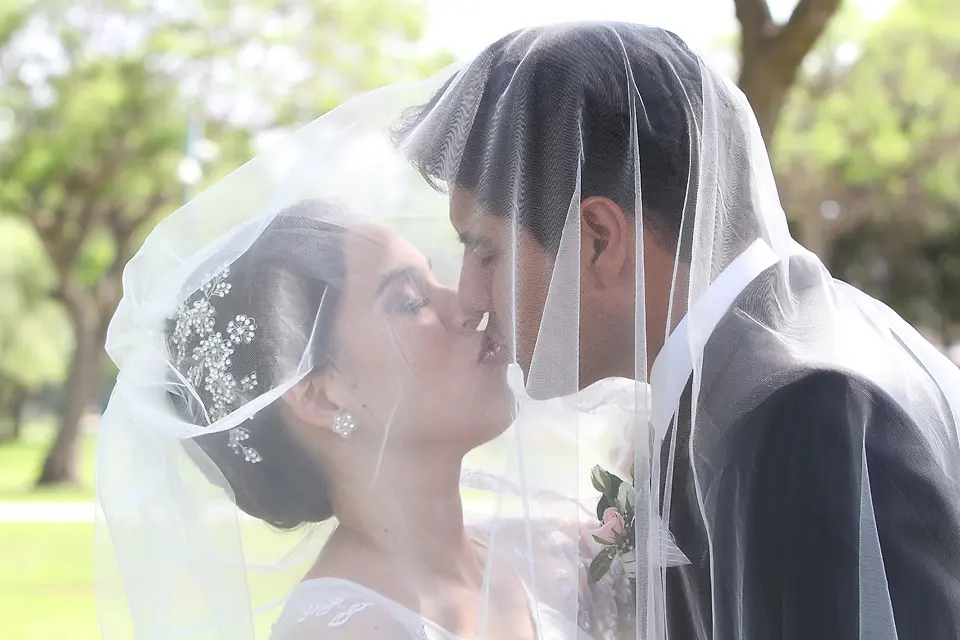 Primer plano íntimo de los novios besándose cubiertos por el velo de la novia en un entorno exterior luminoso.