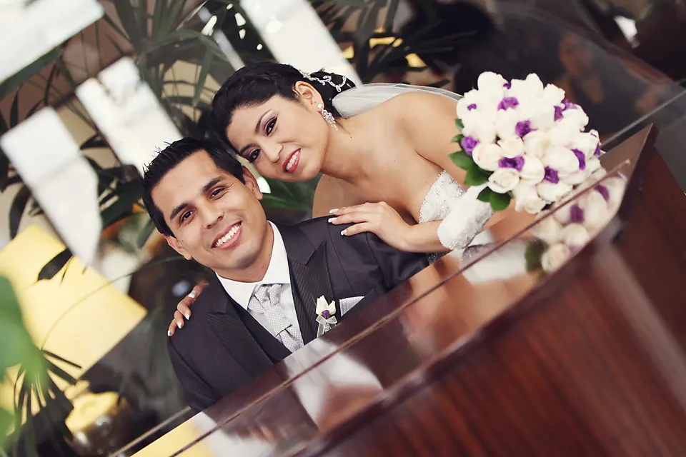 Retrato cercano de los esposos sonriendo; la novia tiene su mano en el hombro del novio y luce un ramo de rosas con detalles morados.