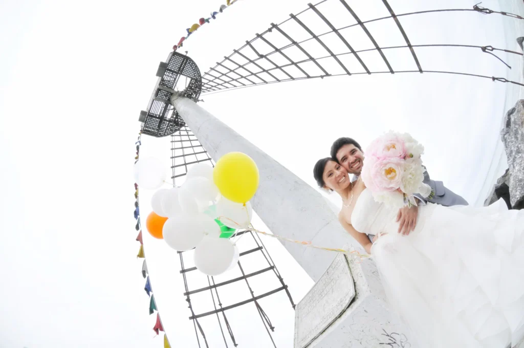 Pareja de novios sonriendo con globos de colores y ramo de novia bajo mástil de barco.
