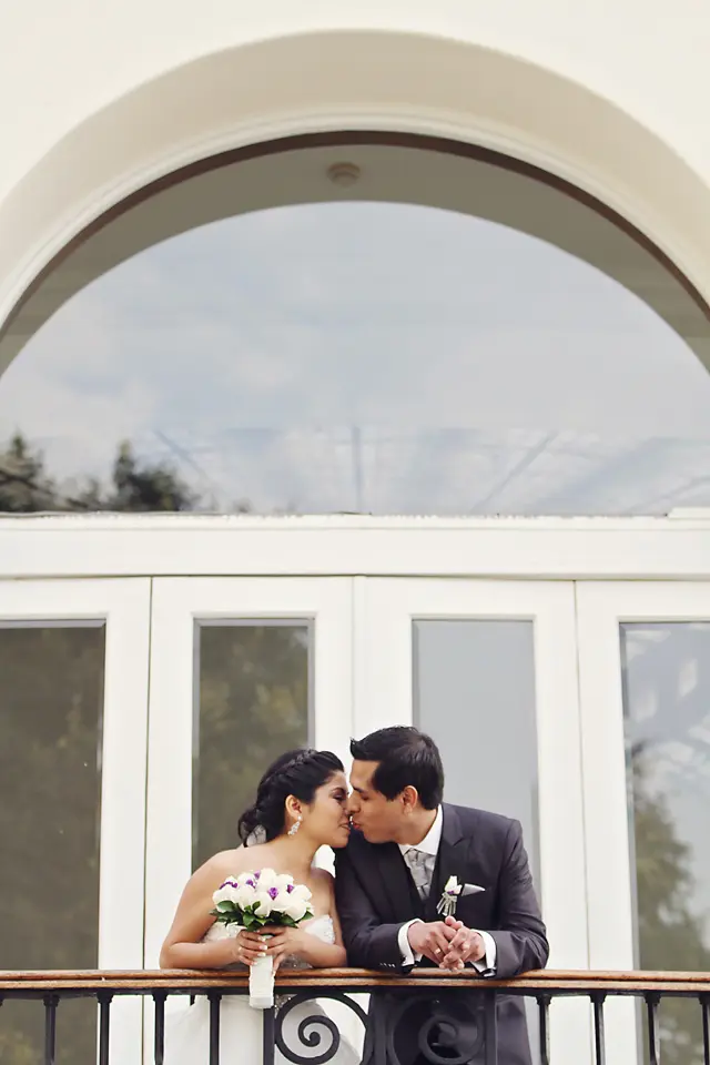 os recién casados se dan un beso romántico apoyados en la baranda de un balcón con diseño de volutas, bajo un gran arco de medio punto.