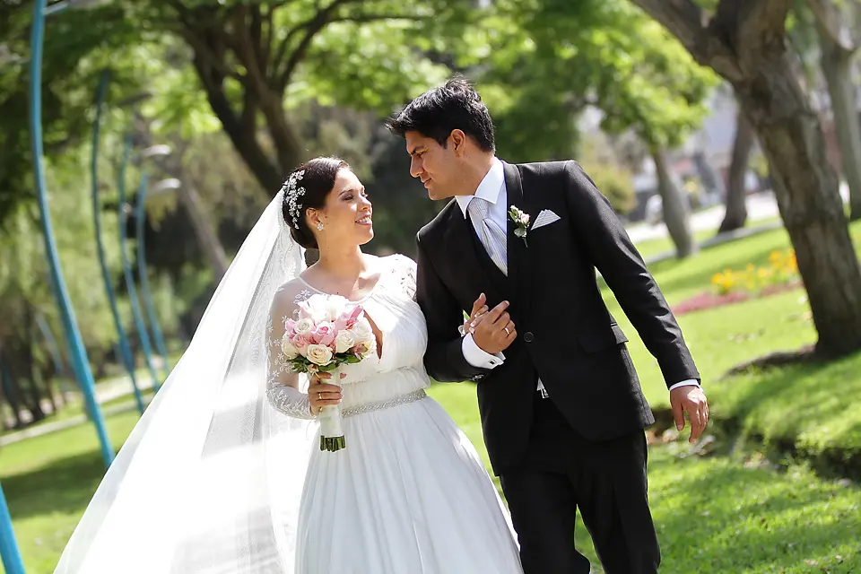 Novios caminando de la mano por un parque verde y sonriéndose mutuamente durante su sesión de fotos de bodas.