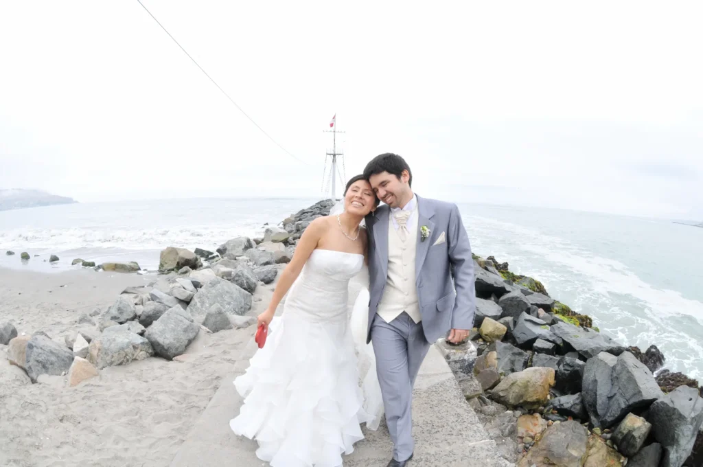 Recién casados caminando y riendo sobre un muelle de piedra frente al mar.