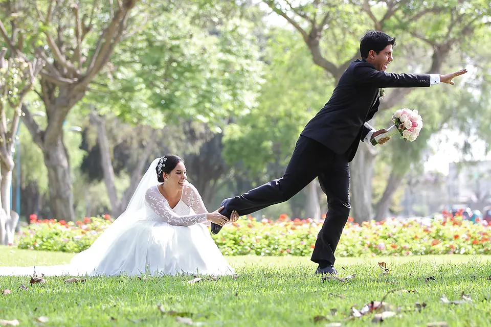 Fotografía divertida de bodas donde la novia en el césped sujeta el pie del novio mientras este intenta escapar sosteniendo el ramo.