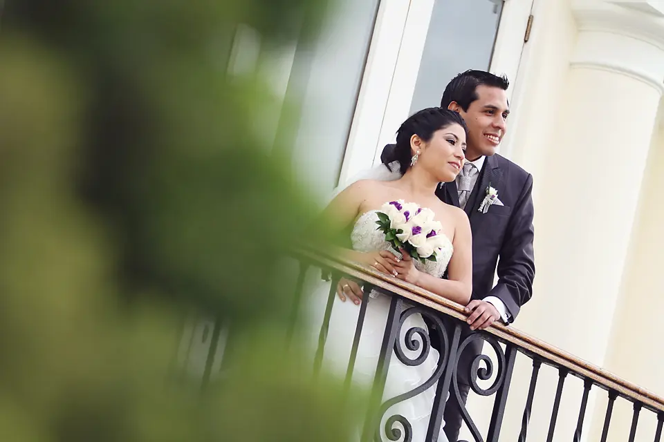 Los novios posan en un balcón clásico; ella sostiene su ramo mientras ambos miran hacia la misma dirección con una expresión serena.