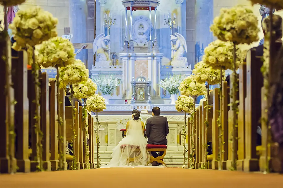 Vista desde el pasillo central de la iglesia hacia el altar, mostrando a los novios arrodillados durante la misa de bodas decorada con flores blancas. Fotografo Bodas Chia