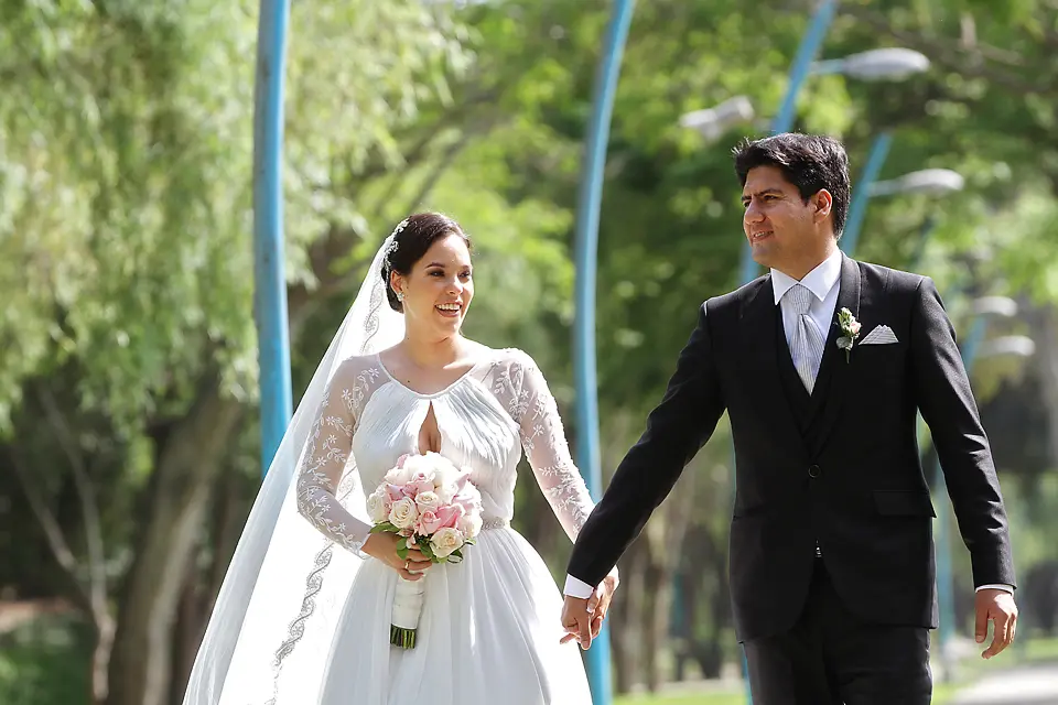 Novios sonrientes caminando de la mano rodeados de árboles durante un día soleado de bodas.