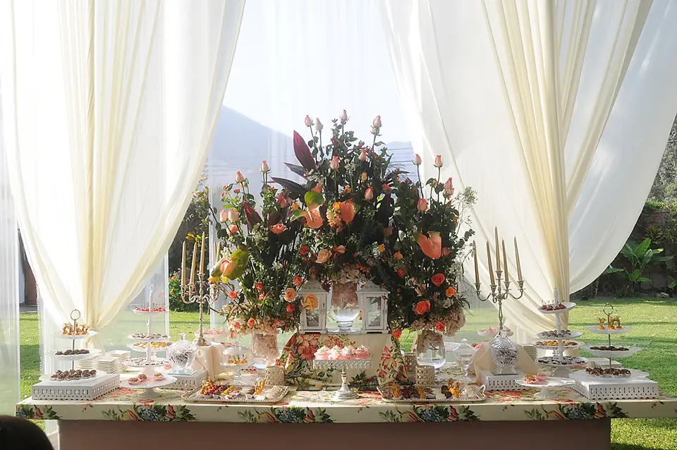 Mesa de postres y dulces de bodas decorada con un gran arreglo floral de rosas, candelabros dorados y cortinas blancas de fondo.