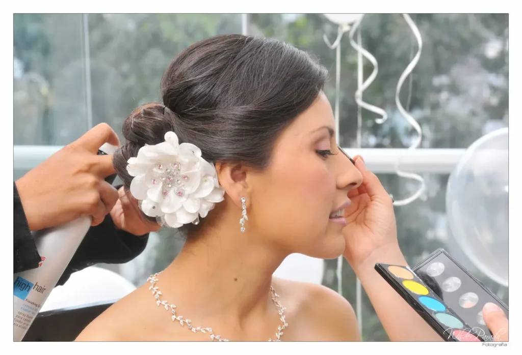 Primer plano lateral de una novia recibiendo laca para el cabello y retoques de maquillaje antes de la boda, destacando un tocado de flor blanca grande en su peinado recogido.