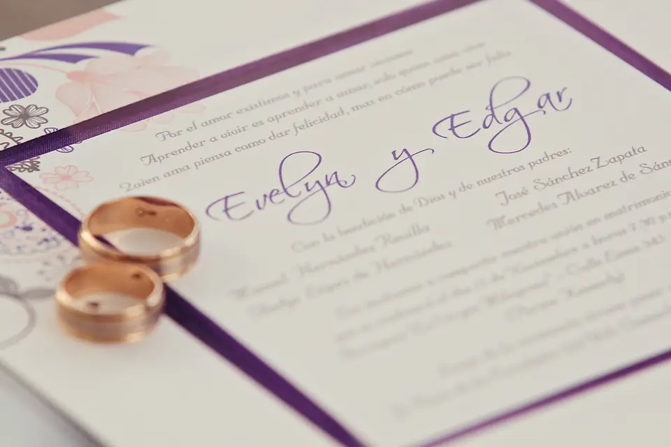 Detalle de una invitación de boda elegante para Evelyn y Edgar con caligrafía morada, acompañada de dos argollas matrimoniales doradas sobre el papel.