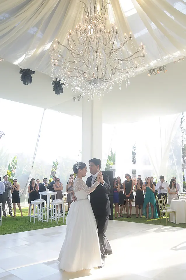 Novios bailando el vals en la pista durante la recepción de su boda, bajo una gran lámpara de cristal elegante y un techo de telas blancas.