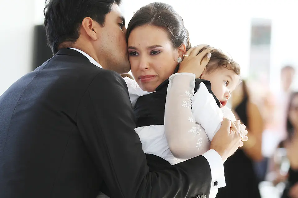 Fotografía conmovedora de la novia llorando de emoción mientras abraza a su pequeño hijo y recibe un beso del novio en la mejilla.