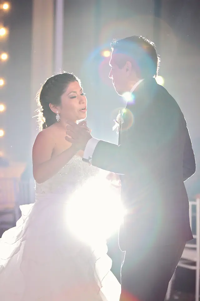 La novia sonríe con complicidad mientras baila con el novio bajo una iluminación cálida, destacando su peinado recogido y pendientes brillantes.