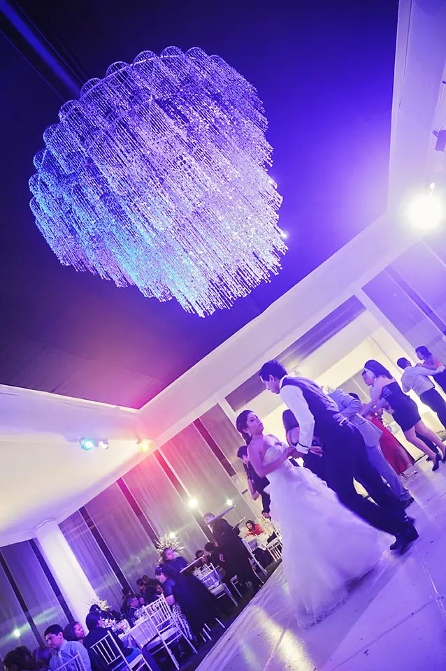 Plano angular de la recepción de la boda mostrando a los invitados en la pista de baile bajo una imponente lámpara de cristal con iluminación azul y violeta.