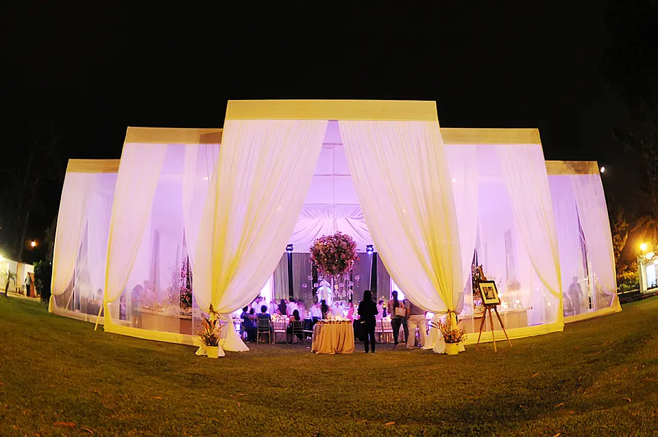 Vista exterior nocturna de una gran carpa de bodas iluminada con luces cálidas y violetas, decorada con elegantes cortinas blancas en un jardín.