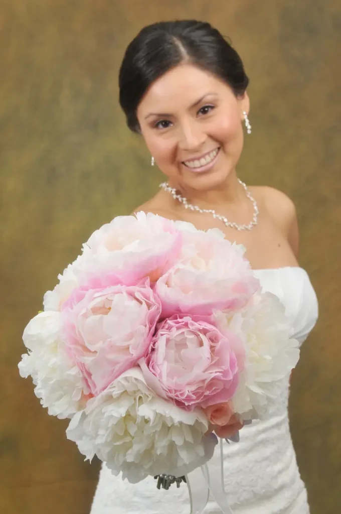 etrato enfocado en un ramo de flores compuesto por peonías blancas y rosas, sostenido por una novia sonriente que aparece levemente desenfocada al fondo.