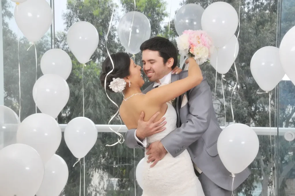 Una pareja de recién casados, ella con vestido de novia blanco y él con traje gris, abrazándose alegremente en una habitación decorada con numerosos globos blancos flotantes.
