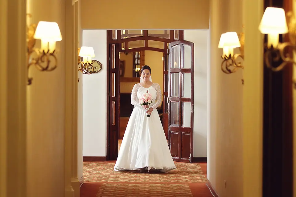 Novia sonriente con vestido blanco y ramo caminando por un pasillo iluminado con lámparas de pared, fotografía de bodas en Bogotá.
