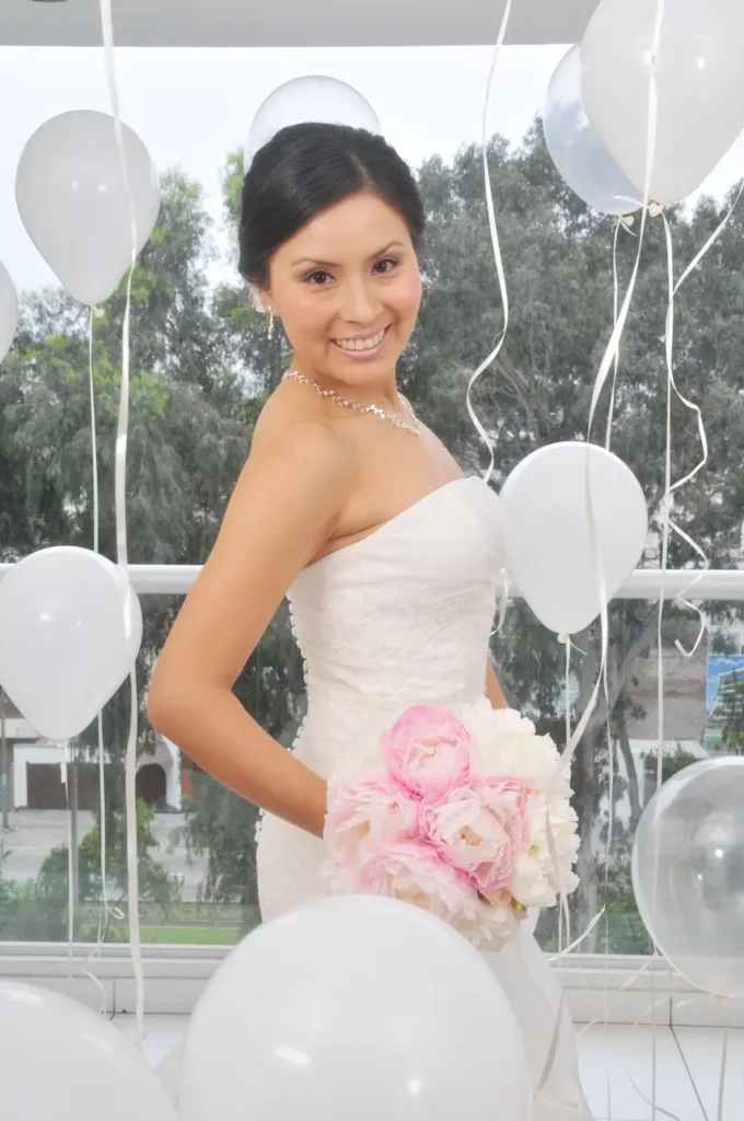 Retrato de una novia de perfil sonriendo a la cámara, sosteniendo un ramo de flores rosadas y blancas, rodeada de globos blancos y cintas decorativas.