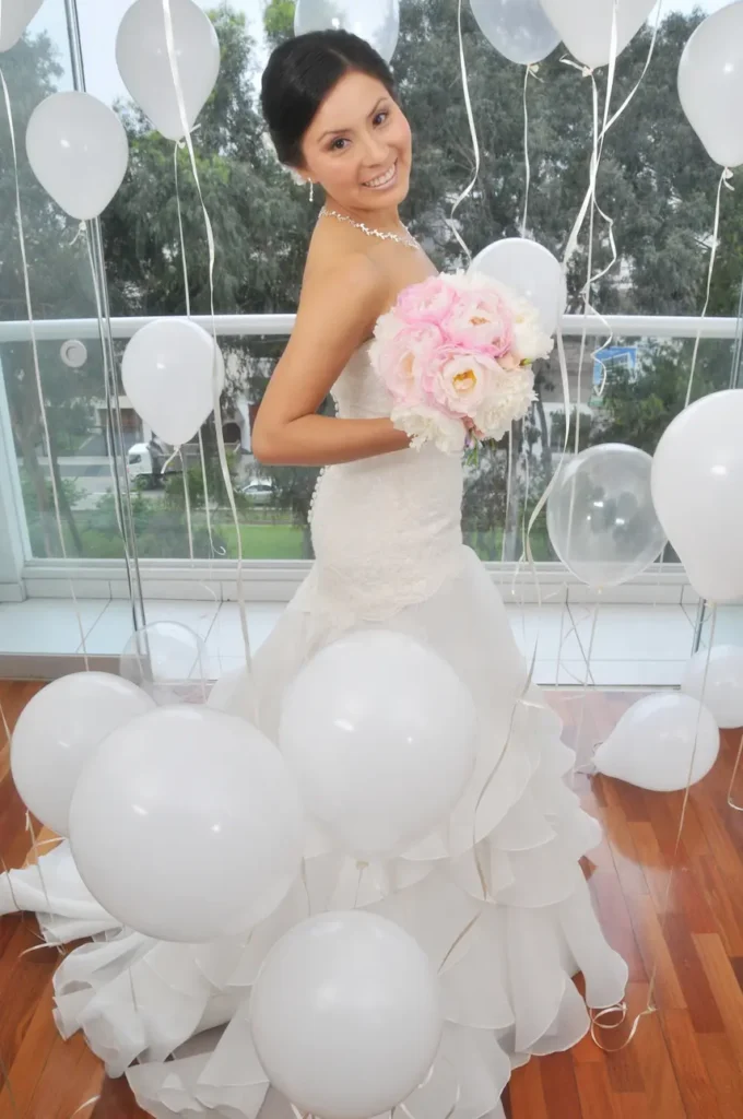 Novia de cuerpo entero con un vestido blanco de volantes, rodeada de globos blancos en el suelo y flotando, sosteniendo un ramo de flores.
