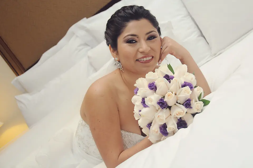 La novia sonríe recostada sobre sábanas blancas sosteniendo un ramo de rosas blancas y flores moradas, luciendo un vestido de novia strapless bordado.