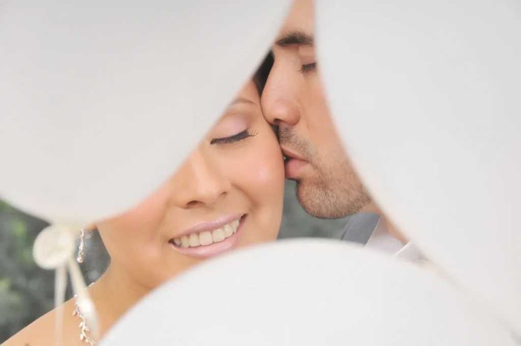 Primer plano extremo de un novio besando la mejilla de la novia, con globos blancos en primer plano creando un efecto de marco desenfocado.