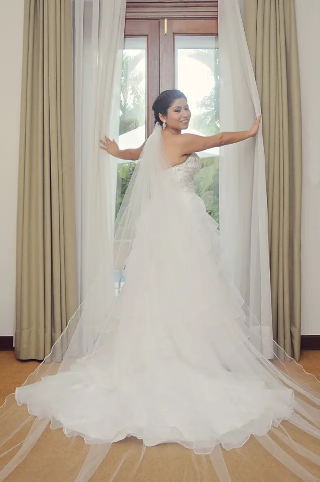 Fotografía de cuerpo completo de la novia de espaldas mirando hacia un ventanal, destacando la larga cola de volantes de su vestido y su velo transparente.