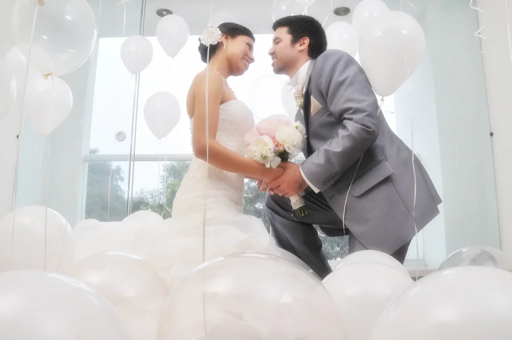 El novio y la novia arrodillados sobre un suelo cubierto de globos blancos, mirándose fijamente mientras sostienen juntos el ramo de flores.