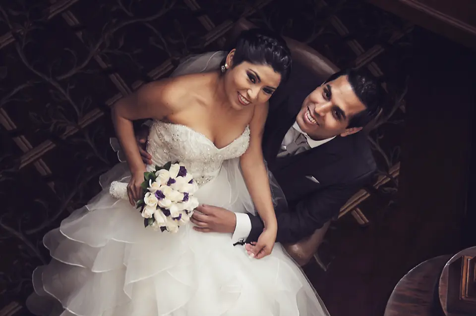 Toma cenital de los novios sentados juntos; ella sostiene un ramo de flores blancas y moradas mientras el novio la abraza con ternura. Wilder Fotografia