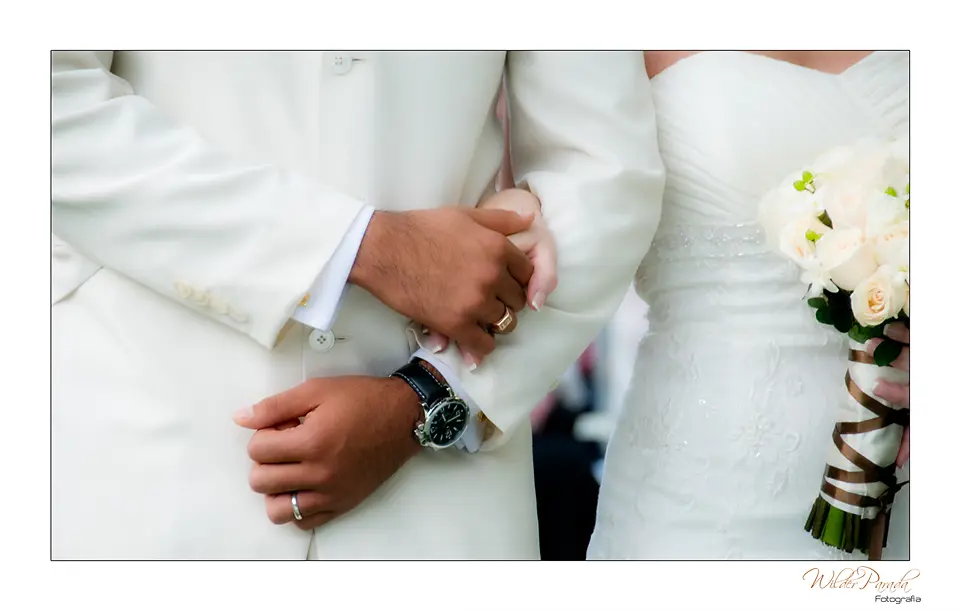 Plano detalle de las manos de los novios, alianzas de matrimonio y ramo de flores blancas en un evento en Bogotá