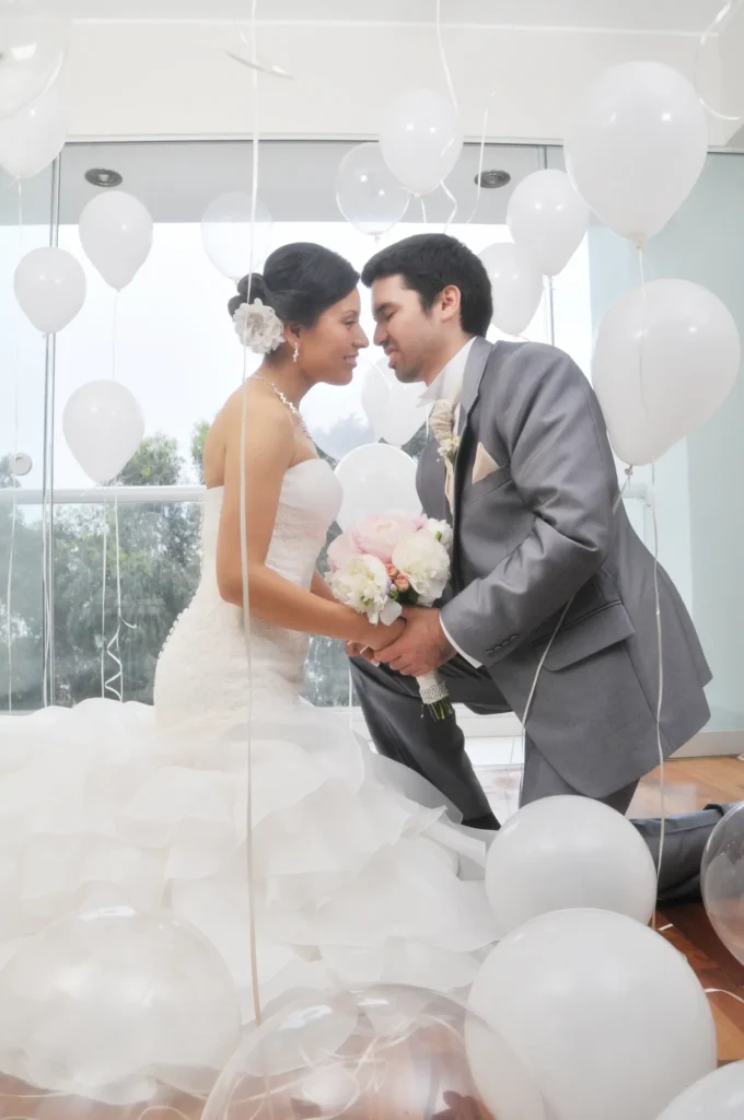 Imagen de los novios de rodillas rodeados de globos blancos; el novio sostiene el ramo mientras ambos se acercan para compartir un momento romántico.