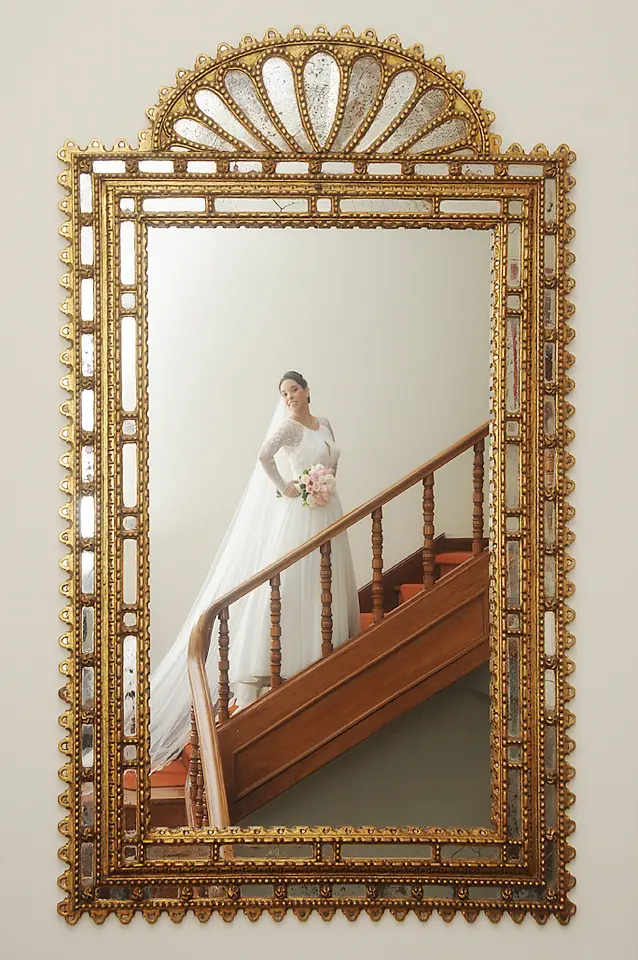 Reflejo en espejo dorado antiguo de una novia con vestido blanco posando en las escaleras, fotografía de bodas en Cundinamarca.